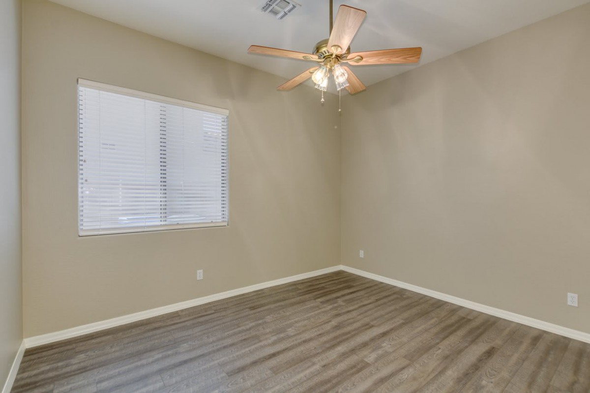 An empty room with hardwood flooring, beige walls, a ceiling fan, and a window with blinds.