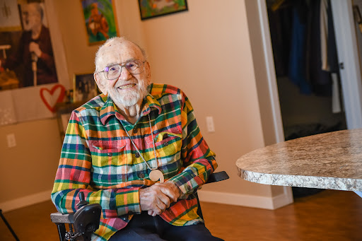 An elderly man with a warm smile sits in a colorful plaid shirt, enjoying a moment indoors.