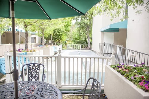 A cozy outdoor patio with a table and chairs under an umbrella, overlooking a fenced area surrounded by trees.