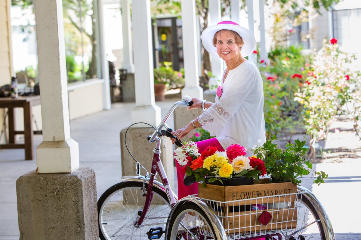 A woman wearing a hat rides a tricycle filled with flowers outdoors on a sunny day.