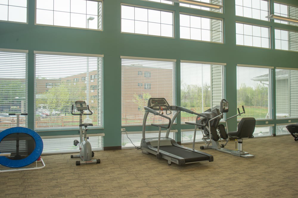 A gym room with various exercise equipment including a treadmill and a stationary bike, set against large windows.