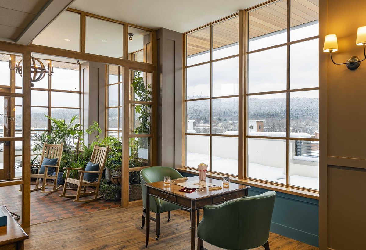 A cozy restaurant interior with large windows providing a scenic view, featuring wooden floors and lush indoor plants.