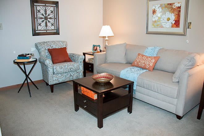 A cozy living room with a beige sofa, patterned armchair, and dark wooden coffee table, accented with colorful cushions and wall art.