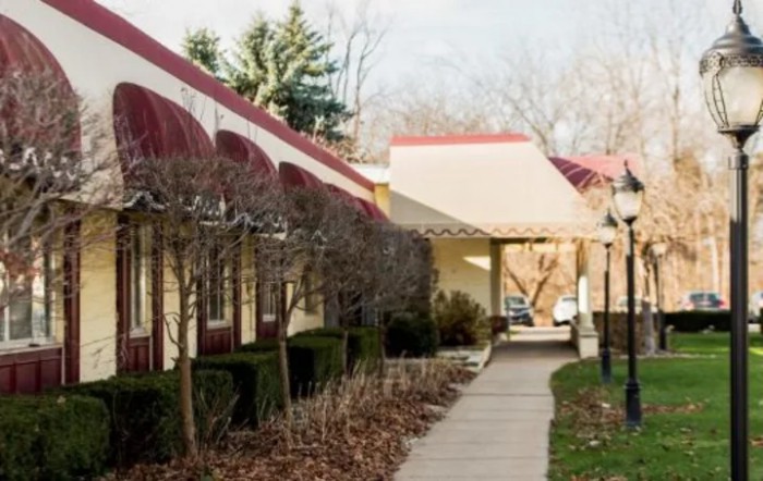 A walkway lined with trimmed bushes and trees, adjacent to a building with red awnings.