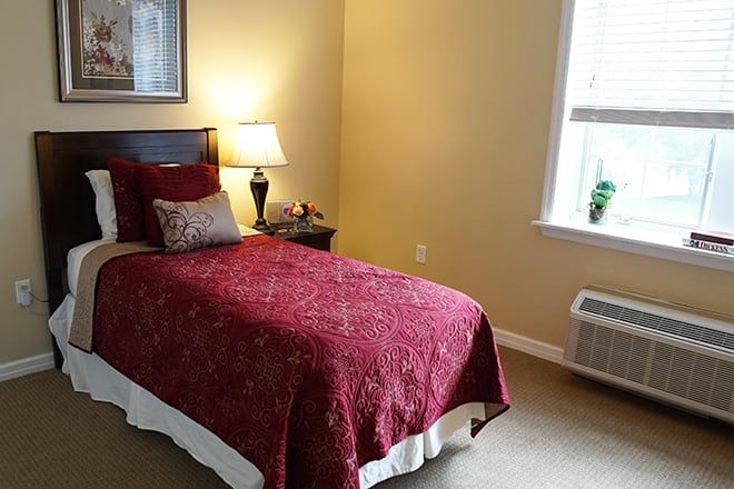 A cozy bedroom featuring a single bed with a red cover, a bedside lamp, and a window allowing natural light.