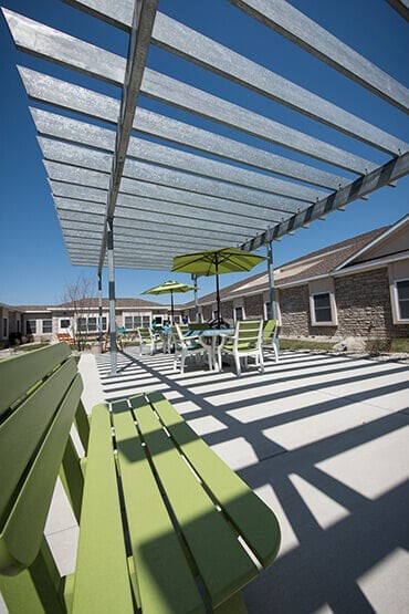 A modern outdoor patio area with green chairs, tables, and umbrellas under a pergola.