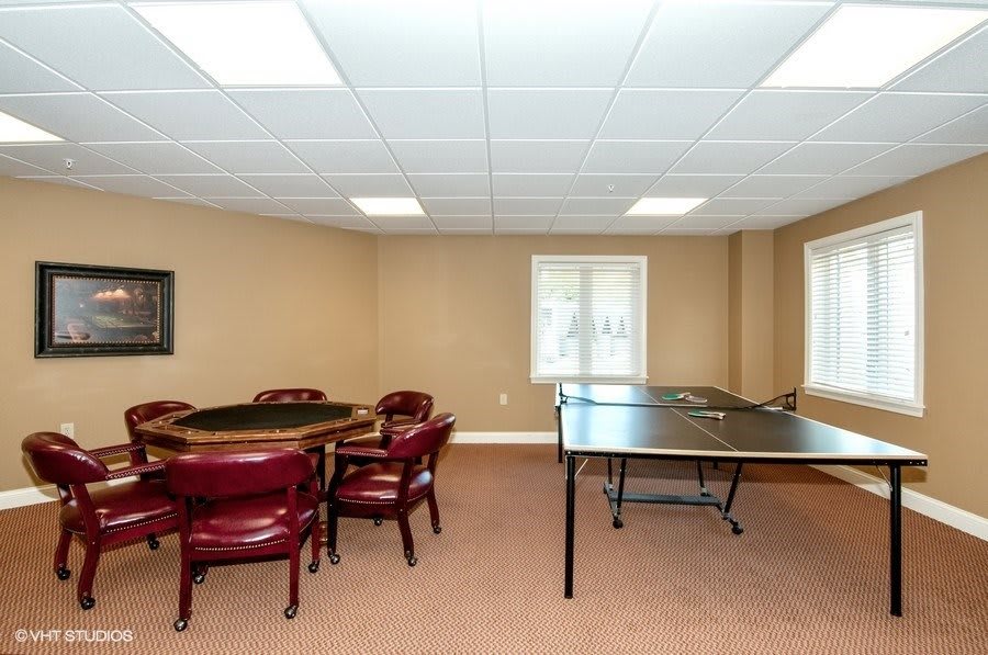 A spacious game room featuring a ping pong table and a card table with chairs, complemented by carpeted flooring and bright overhead lighting.
