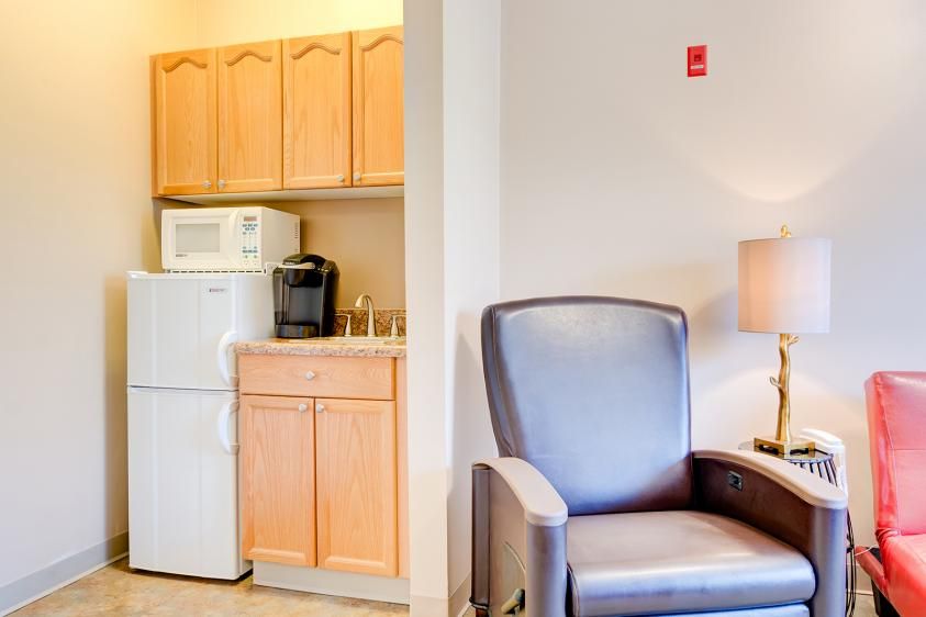 A cozy interior space featuring a small kitchenette with a refrigerator, microwave, and coffee maker next to a comfortable armchair and lamp.