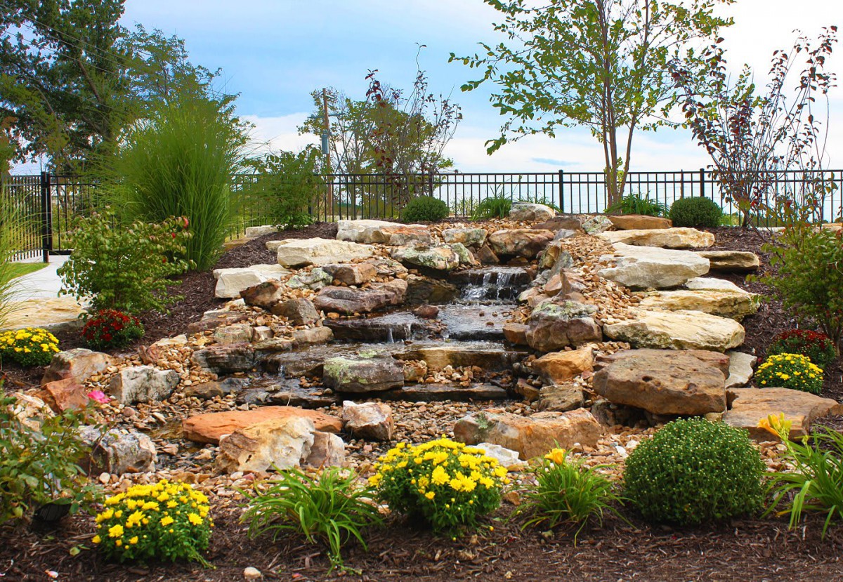 A landscaped garden featuring a small waterfall cascading over rocks, surrounded by lush greenery and vibrant flowers.