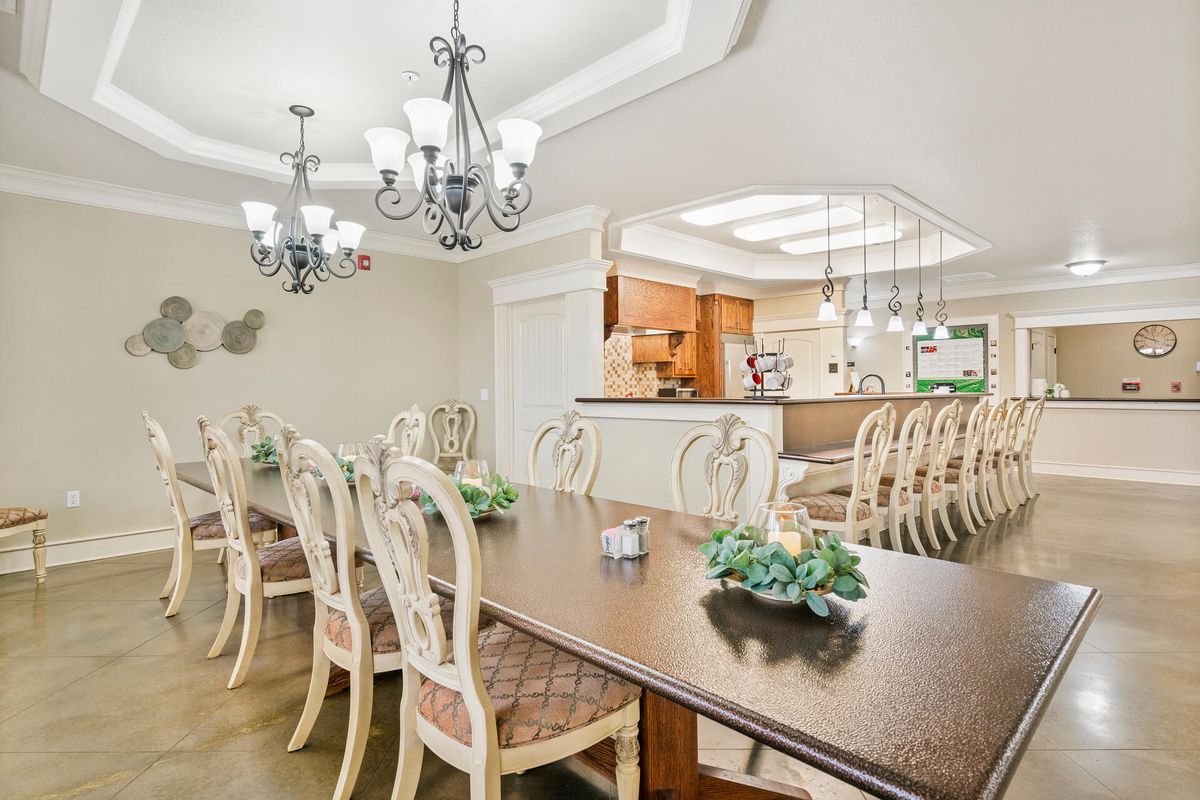 An elegant dining room with a long table and multiple chandeliers, overlooking a kitchen.