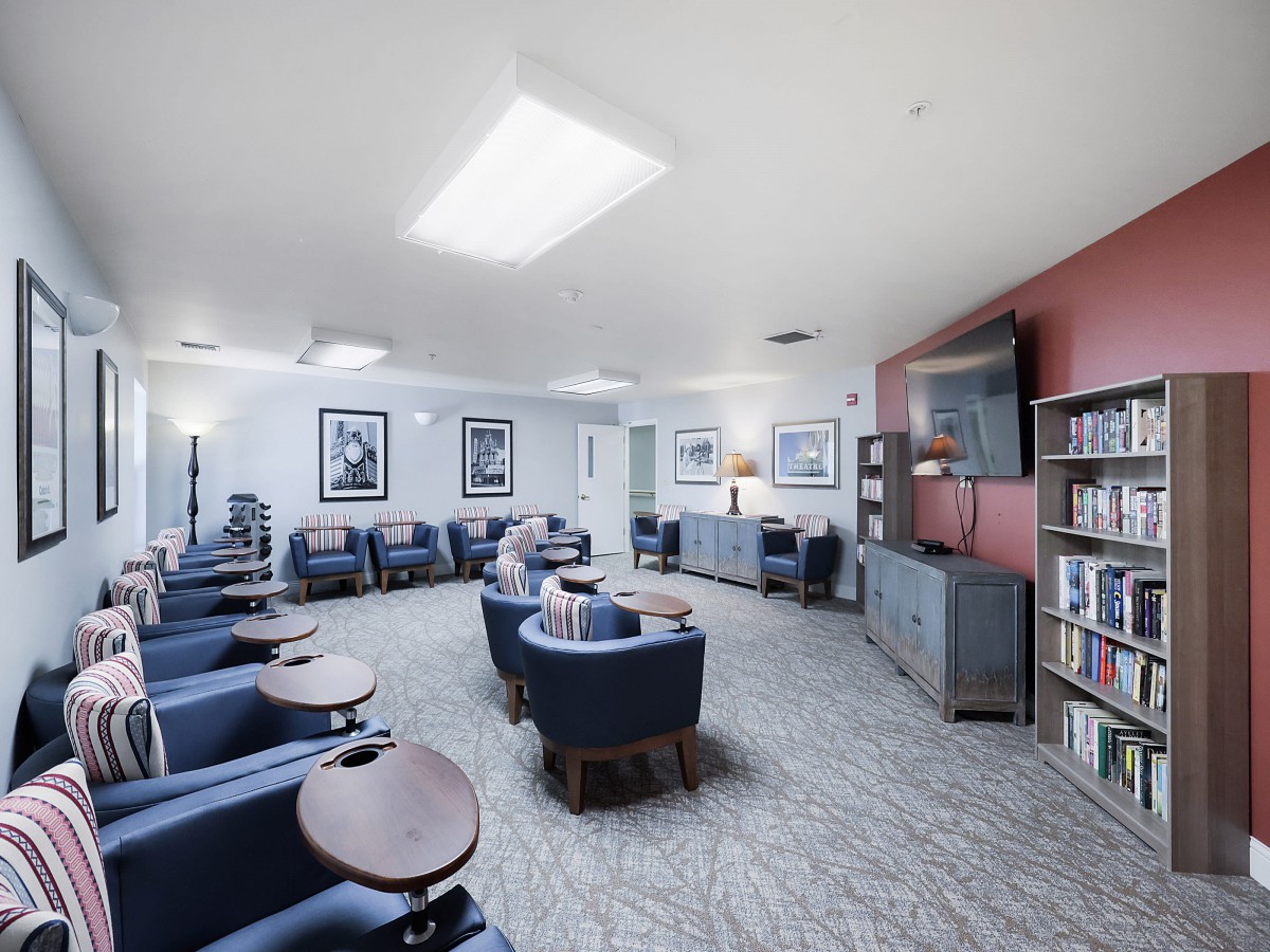 A spacious lounge area with comfortable seating, featuring individual chairs and small tables, a television, and bookshelves lining the walls.