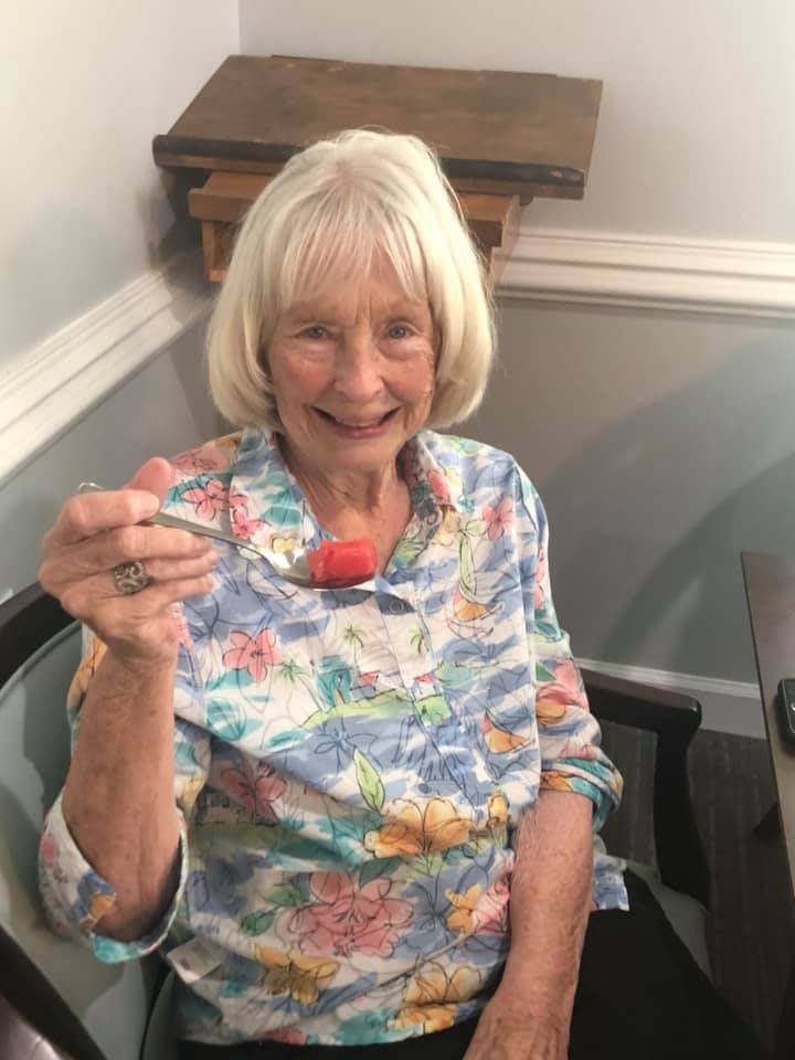 An elderly woman smiling while holding a spoon in her hand indoors.