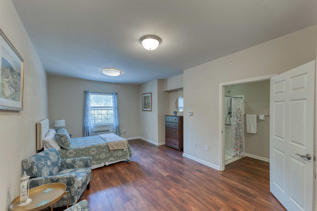 A cozy bedroom with a wooden floor, featuring a single bed, armchair, and an adjoining bathroom.