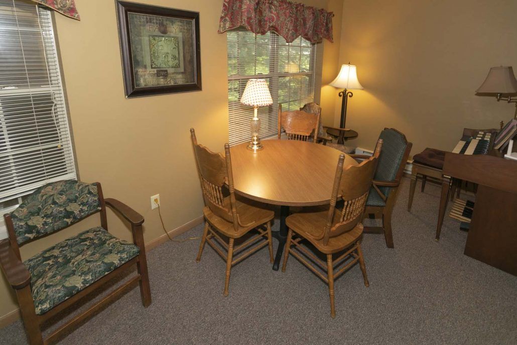 A cozy dining area with a round wooden table surrounded by chairs, softly lit by a lamp near the window.