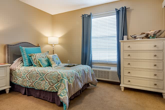 A cozy bedroom with a neatly made bed and colorful cushions, featuring a dresser and a window with blue curtains.