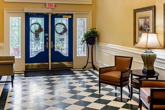 A lobby with checkered flooring, featuring a single chair, a potted plant, and blue double doors adorned with wreaths.