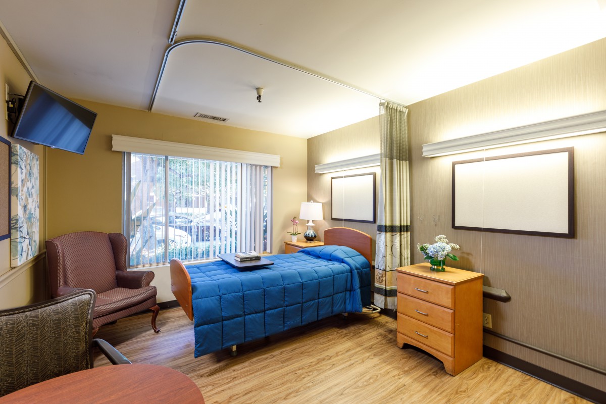 A hospital room with a single patient bed, wooden furniture, and a window with vertical blinds.
