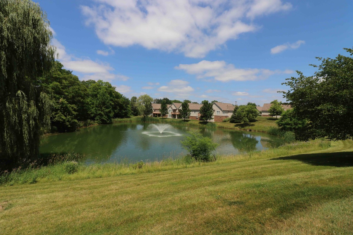 A scenic view of a pond with a fountain, surrounded by grassy slopes and apartment buildings under a clear blue sky.