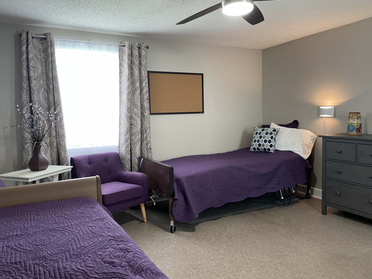 A shared bedroom with two beds adorned in purple bedding, a comfortable armchair by the window, and a bulletin board on the wall.