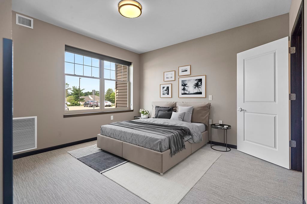 A modern bedroom with neutral tones featuring a bed, rug, and decorative wall art, illuminated by natural light from a large window.