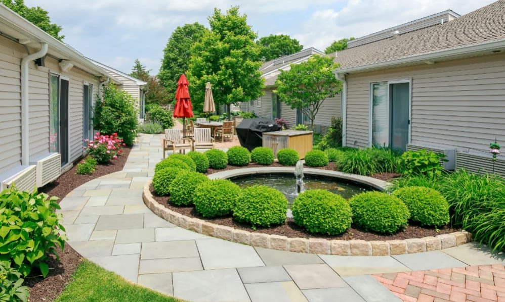 A beautifully landscaped backyard with a circular fountain at the center, surrounded by neatly trimmed shrubbery and stone pathways leading to a cozy patio area.