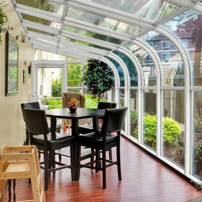 A bright sunroom with a glass ceiling and modern dining furniture, filled with natural light and surrounded by lush greenery.