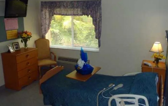 A hospital room featuring a bed, a chair, and a window with blinds and curtains. A piece of towel art is placed on the table over the bed.