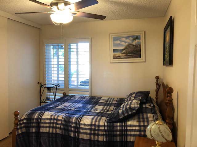 A cozy bedroom with plaid bedding and a ceiling fan, featuring window shutters and artwork on the wall.