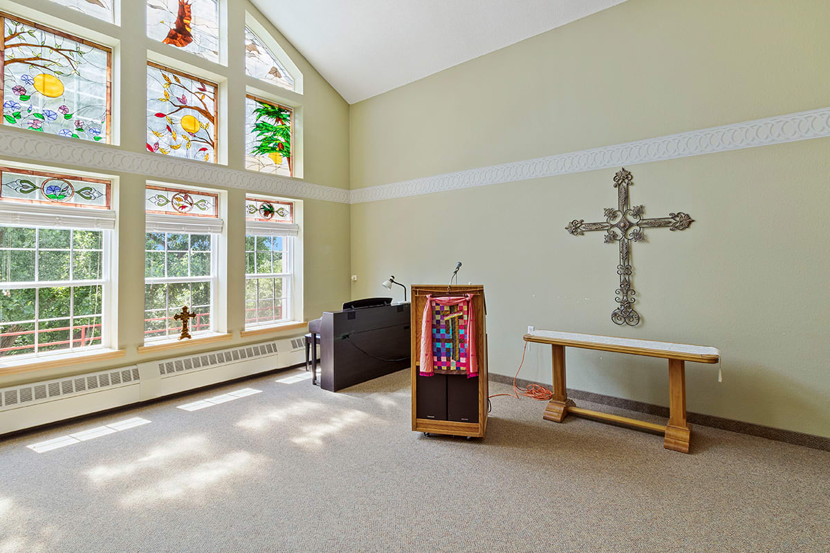 A serene chapel interior with stained glass windows, a cross on the wall, and a wooden pulpit and altar.