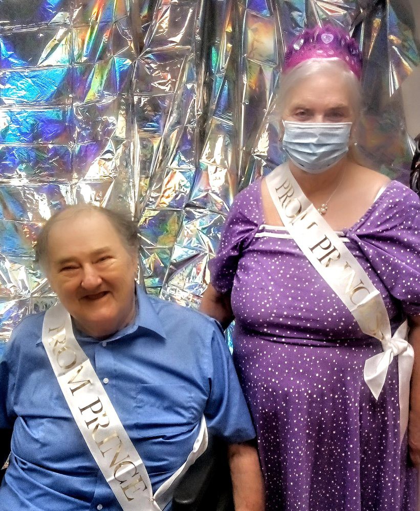 A joyful celebration with two seniors wearing sashes, one in a purple dress and mask, posing against a shiny backdrop.