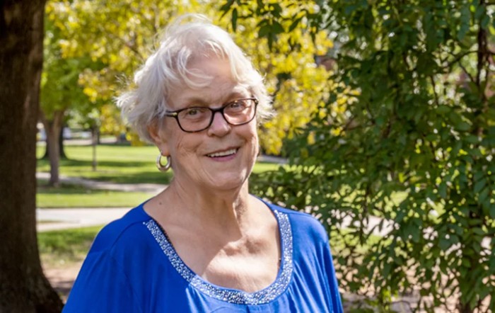 An elderly woman with glasses, smiling outdoors in a blue top surrounded by trees.