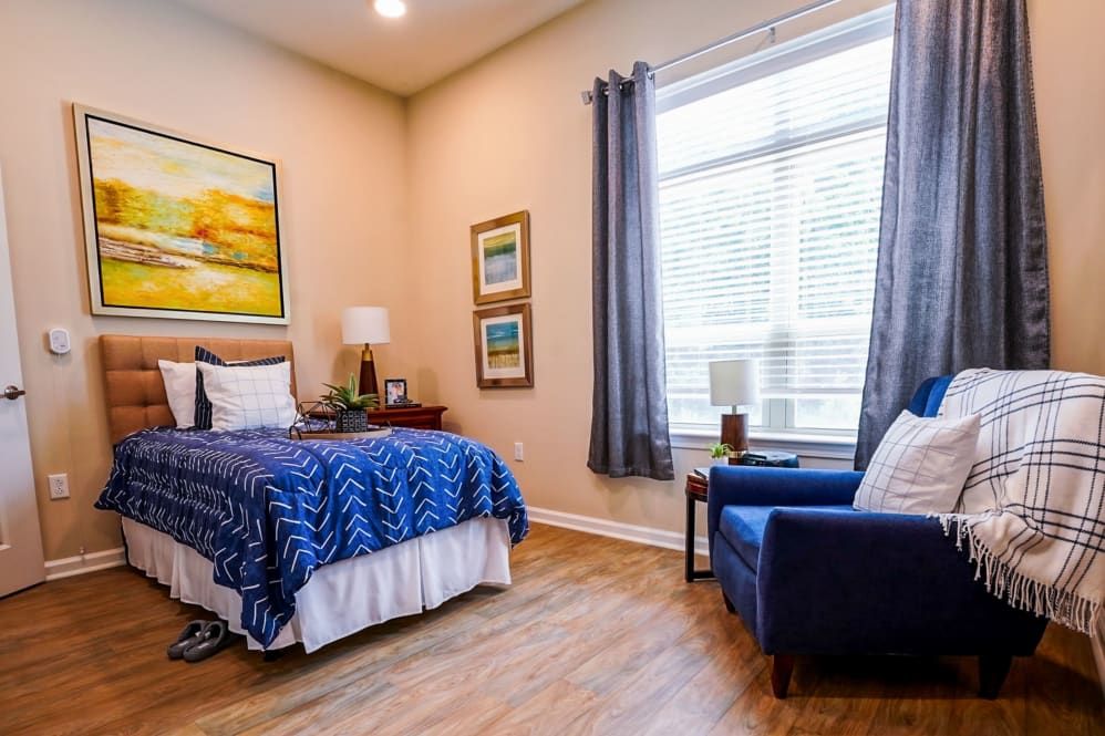 A cozy bedroom with a neatly made bed, art on the walls, and a comfortable chair by the window.