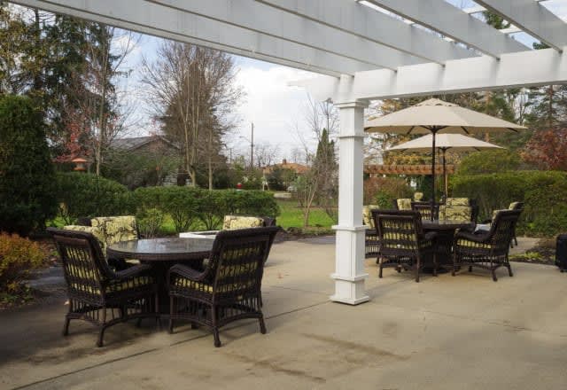 A serene outdoor patio area with comfortable seating under a pergola, surrounded by lush greenery.