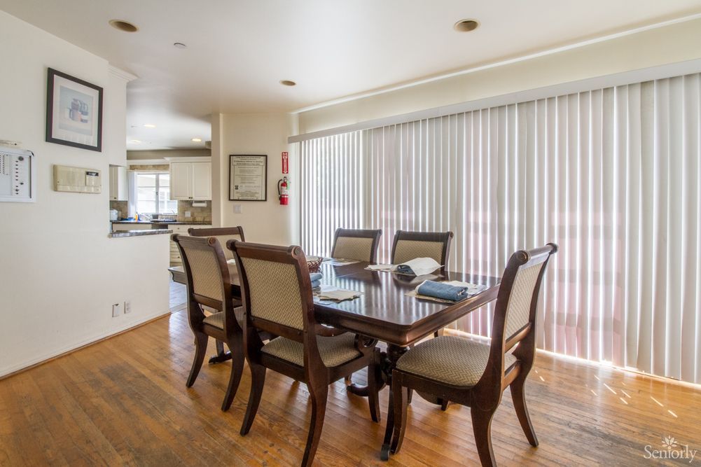 A modern dining room with a glass-topped table and six chairs, featuring sliding doors with vertical blinds and a hardwood floor.