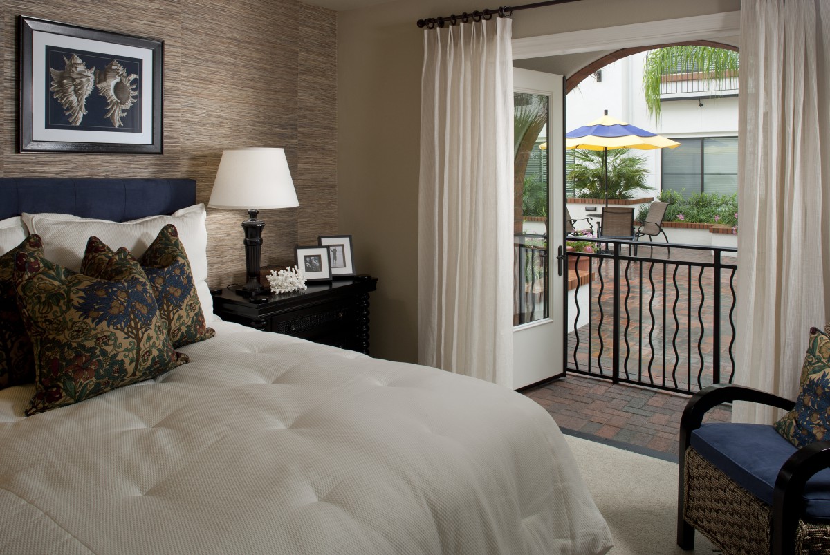 A cozy bedroom with a neatly made bed, nightstand with lamps, and an open door leading to a balcony.