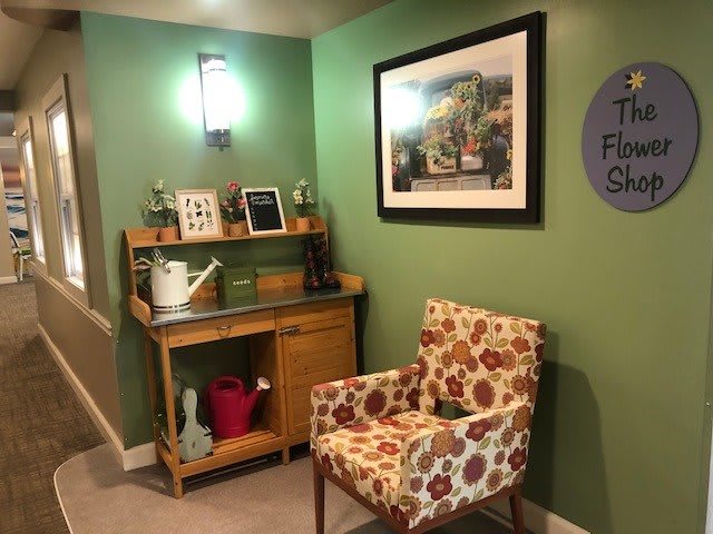 A cozy corner with green walls, featuring floral-patterned chair and wooden desk, complemented by artwork and decorative items.