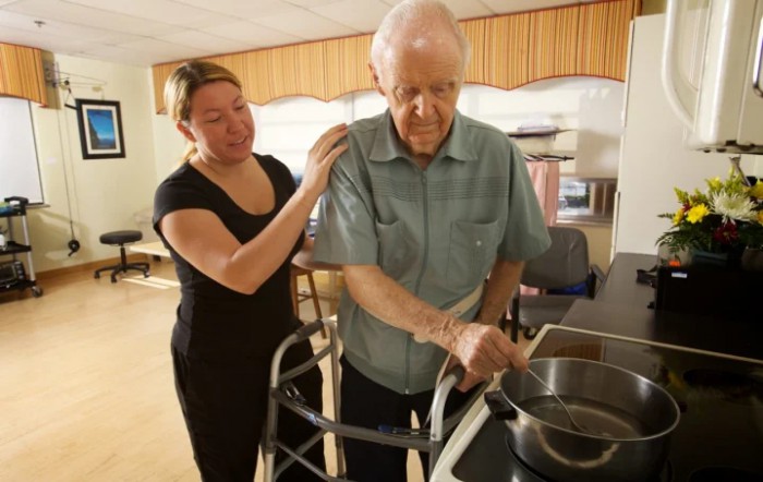 A caregiver assists an elderly man using a walker in the kitchen.