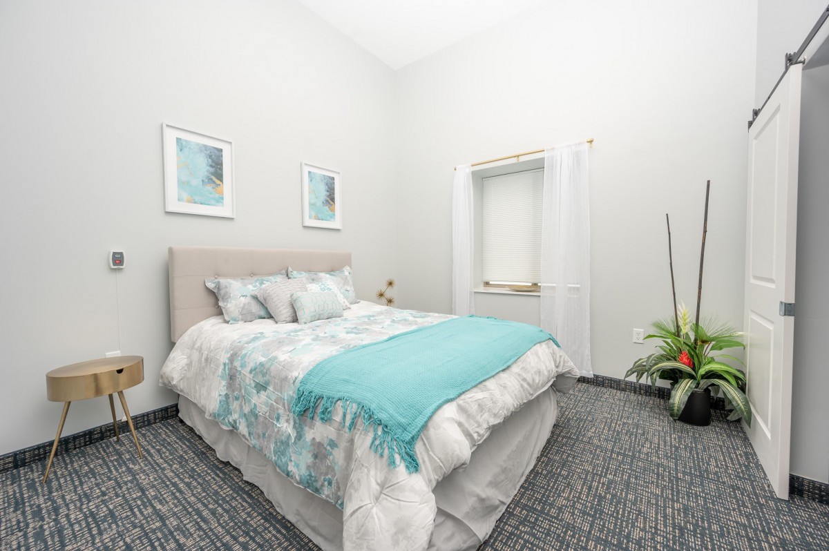 A cozy bedroom with a light color scheme, featuring a neatly made bed, framed artwork on the walls, and decorative plants.