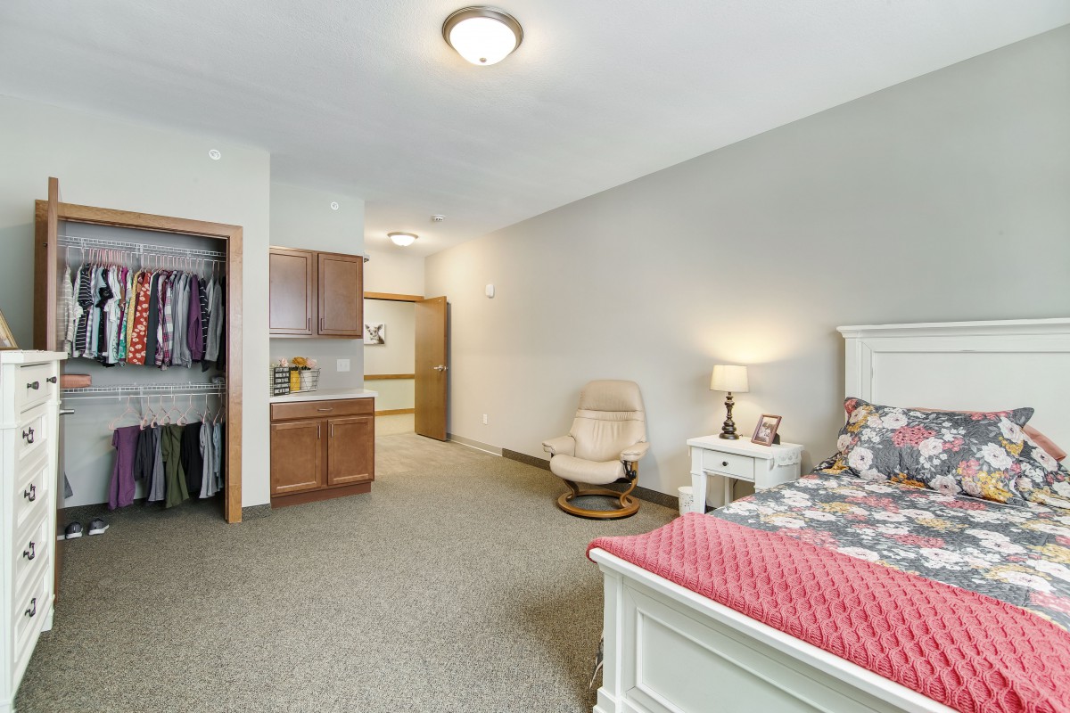 A cozy bedroom with a bed, chair, and lamp, featuring neutral colors and carpet flooring.
