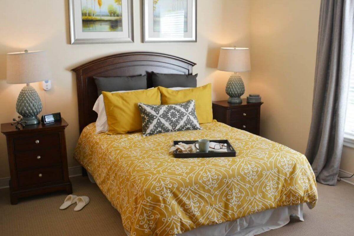 A cozy bedroom with a warm yellow and gray color scheme, featuring a neatly made bed, decorative pillows, and a bedside lamp.