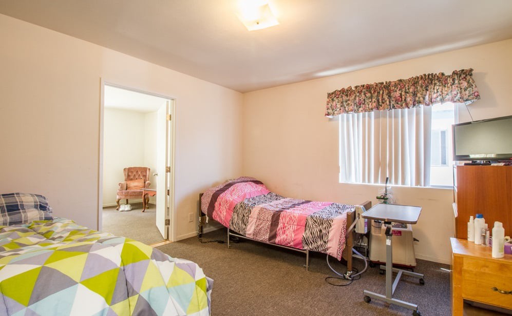 A shared bedroom in a nursing home or hospital with two beds, a small table, and soft lighting.