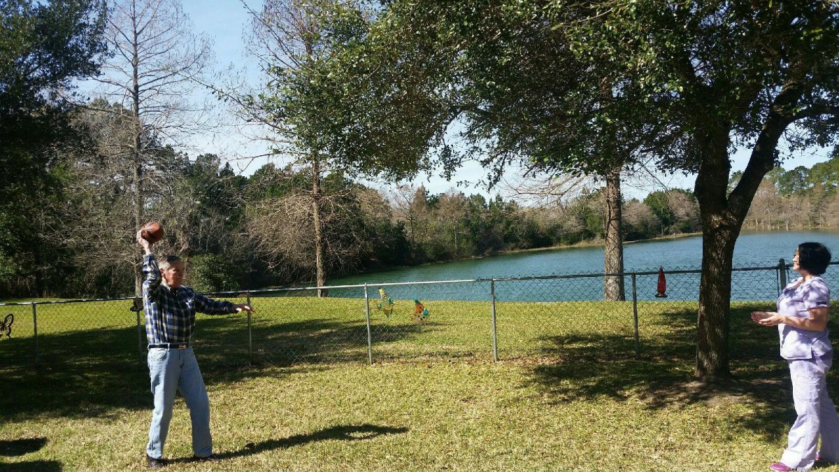 A person standing in a grassy area near a lake, surrounded by trees and a fence.
