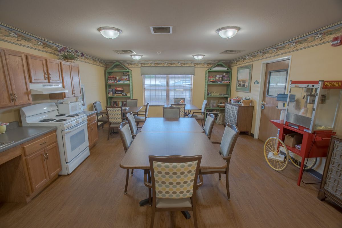 A spacious dining room with a large table surrounded by chairs, featuring kitchen appliances and bookshelves.