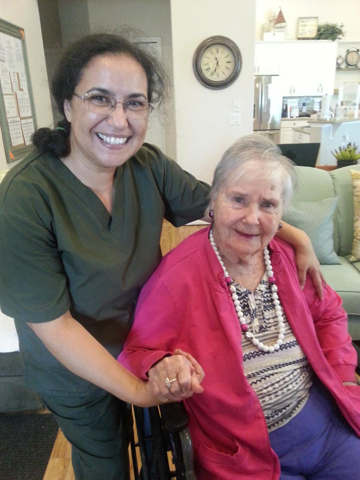 An elderly woman in a pink sweater sits in a wheelchair, holding hands with a smiling caretaker.
