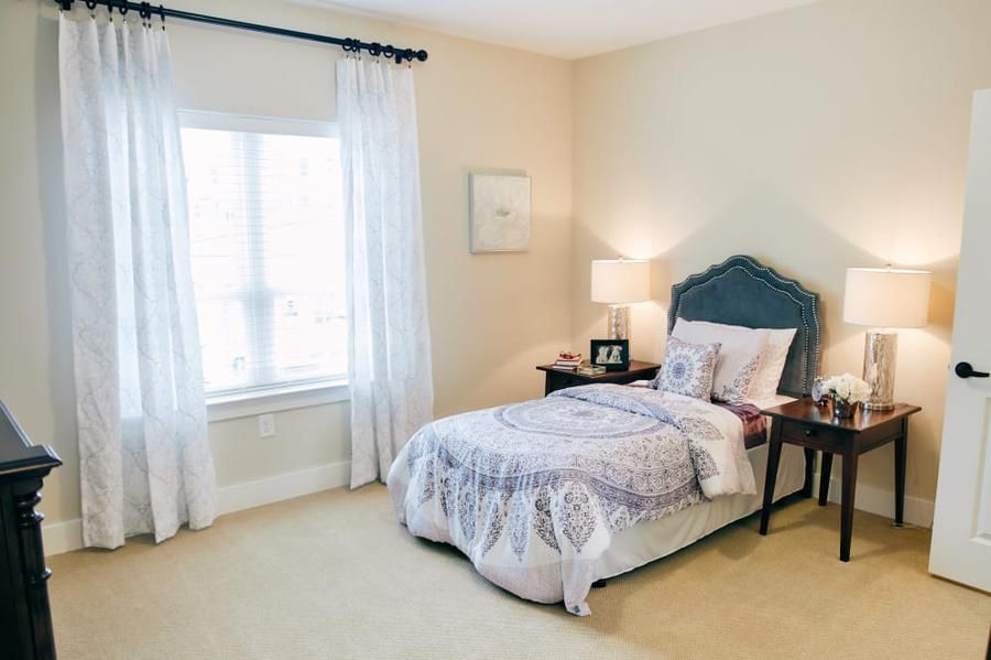 A cozy bedroom with a neatly made single bed beside a window, complemented by curtains and a nightstand with a lamp.