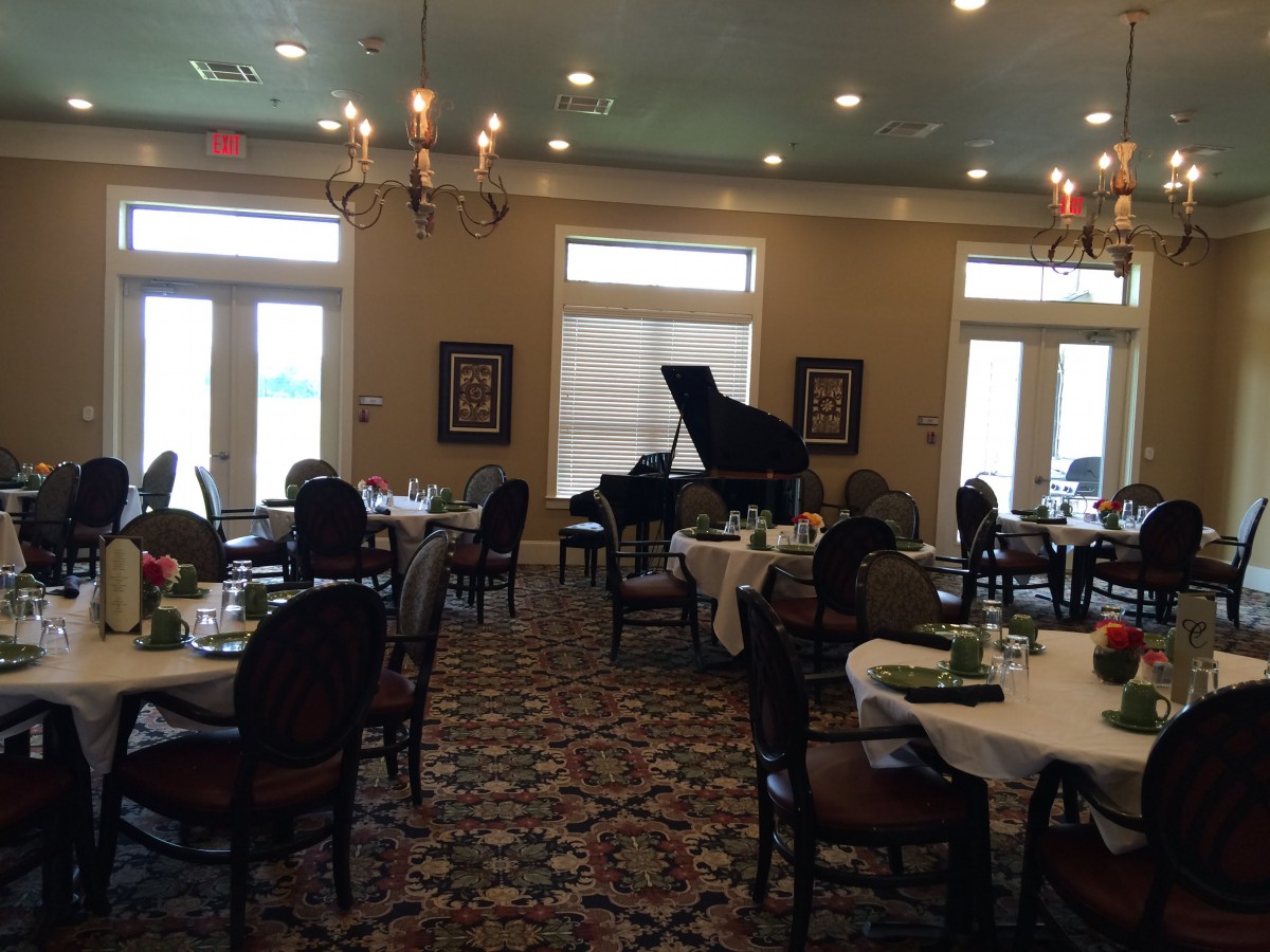 A dining room with round tables, a grand piano, and elegant chandeliers.