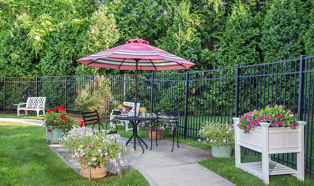 A cozy garden patio setup with a striped umbrella, table, and chairs surrounded by lush greenery and flowers.
