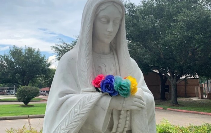 A statue of the Virgin Mary holding colorful flowers and a rosary, set outdoors against a natural backdrop.