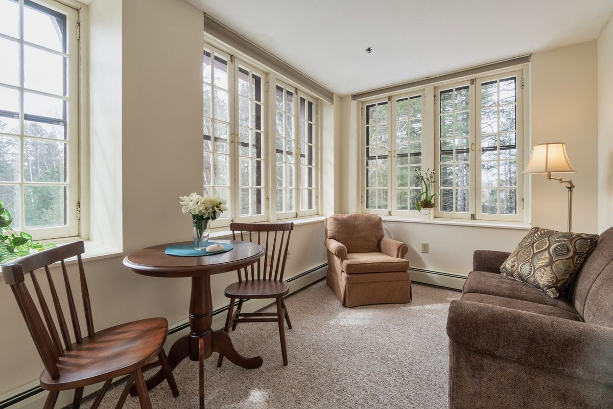 A cozy living room featuring large windows, allowing for ample natural light, with comfortable furniture including an armchair, a small round table, and chairs.