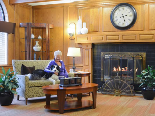 A person sitting on a couch with a dog in a cozy living room featuring a fireplace and large clock.